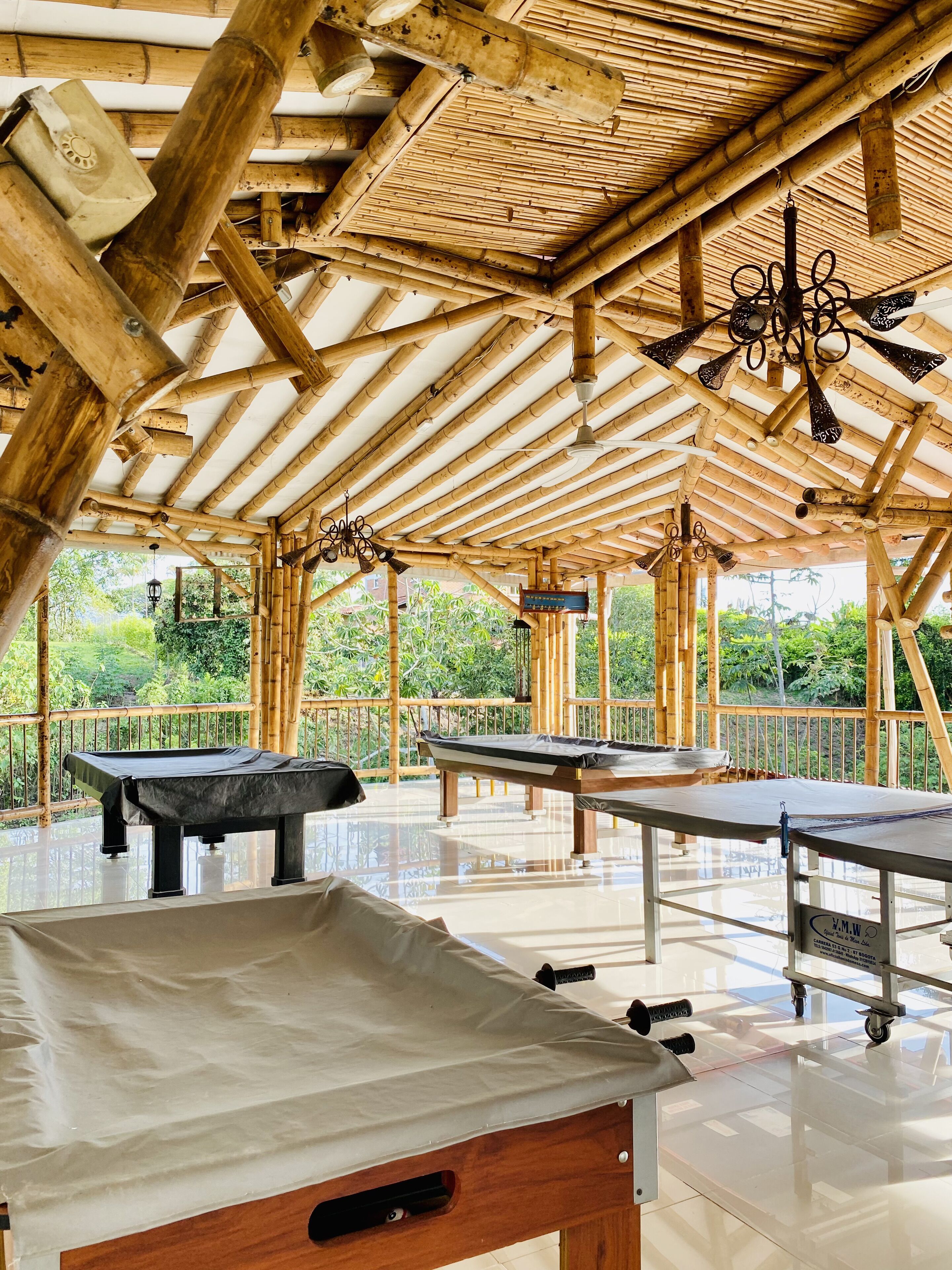 An indoor pool area with bamboo ceiling and walls, multiple pool tables, and a clear view of greenery outside.