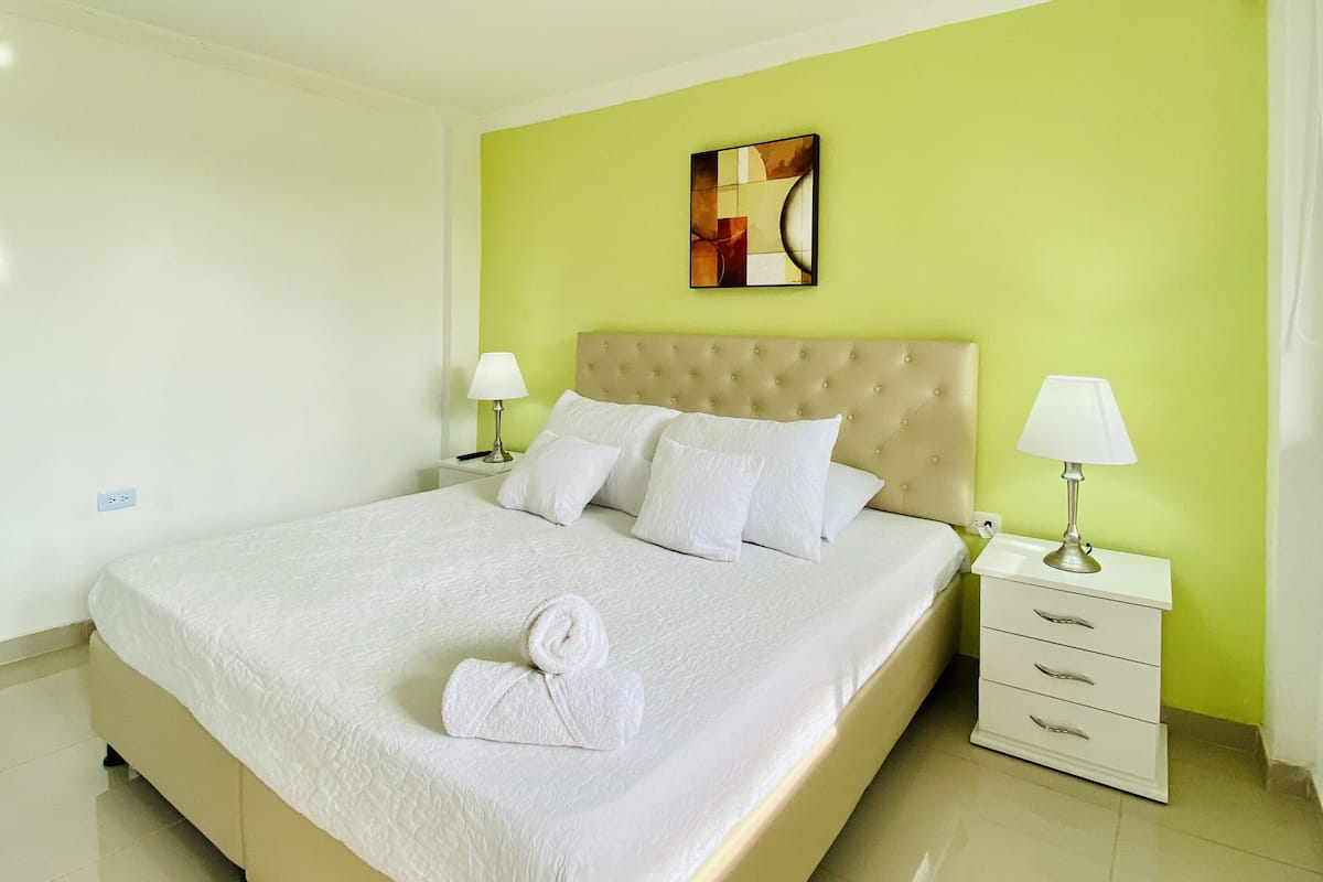 A neatly made bed with white linens and a rolled towel, a tufted headboard, and a bedside table with a lamp.