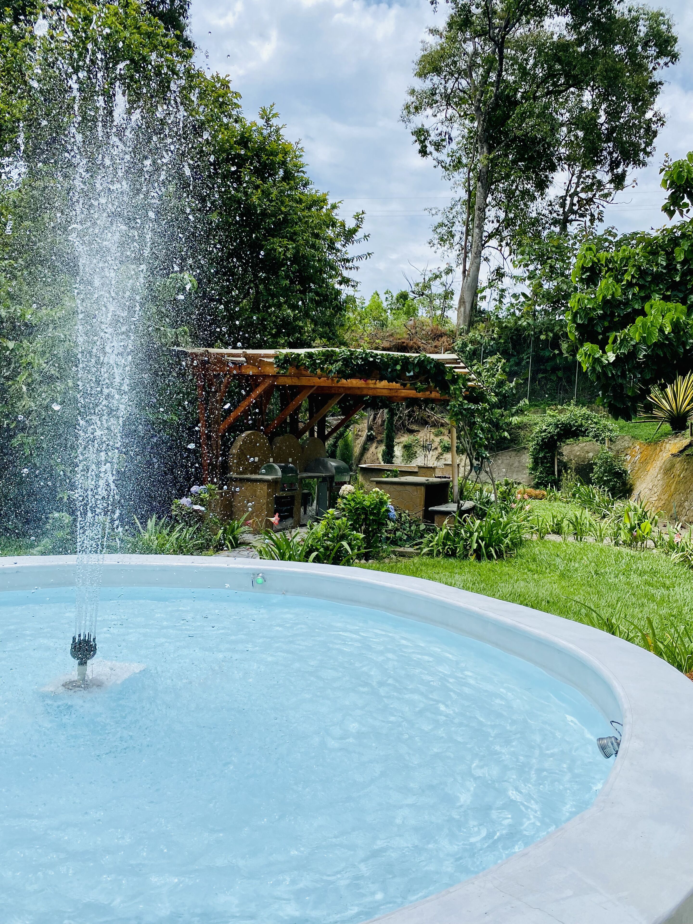 A fountain with water jets in a circular pool, surrounded by lush greenery and a wooden pergola structure.