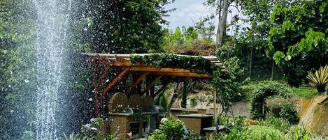 A fountain with water jets in a circular pool, surrounded by lush greenery and a wooden pergola structure.