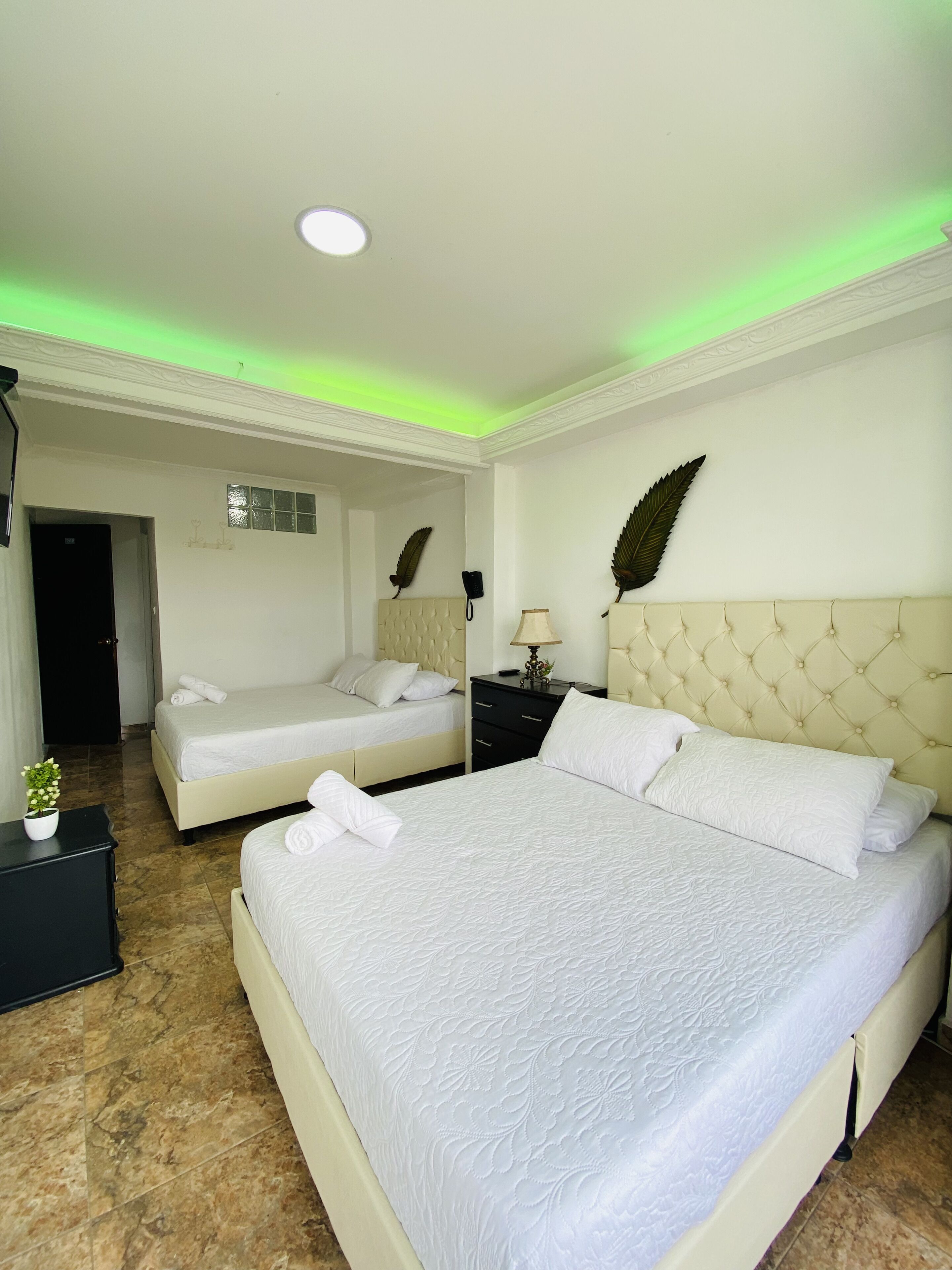 Quarto de hotel com duas camas, uma cama grande com cabeceira estofada e uma cama menor com cabeceira. Há uma mesa de cabeceira com uma luminária e uma folha decorativa na parede.