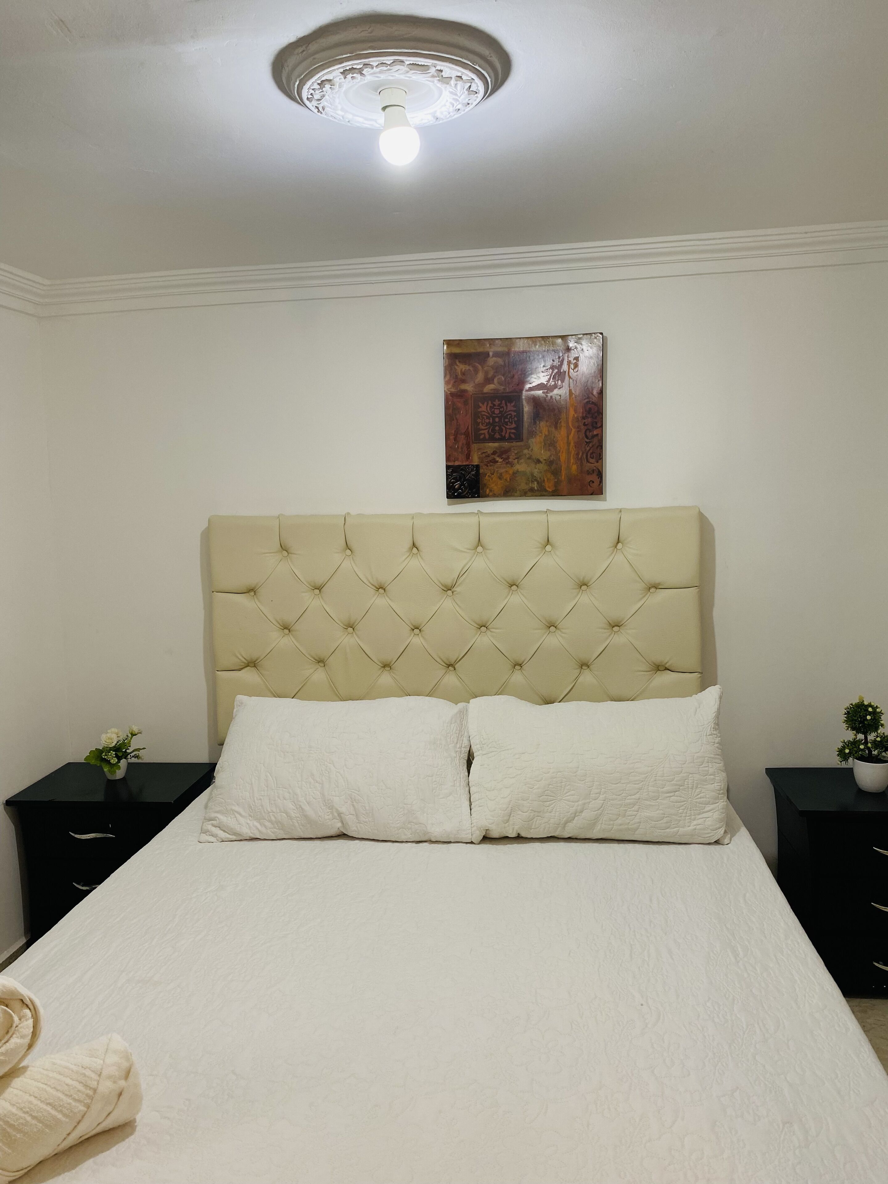 A bedroom with a tufted headboard, a white bedspread, two bedside tables with lamps, and a wall-mounted artwork.