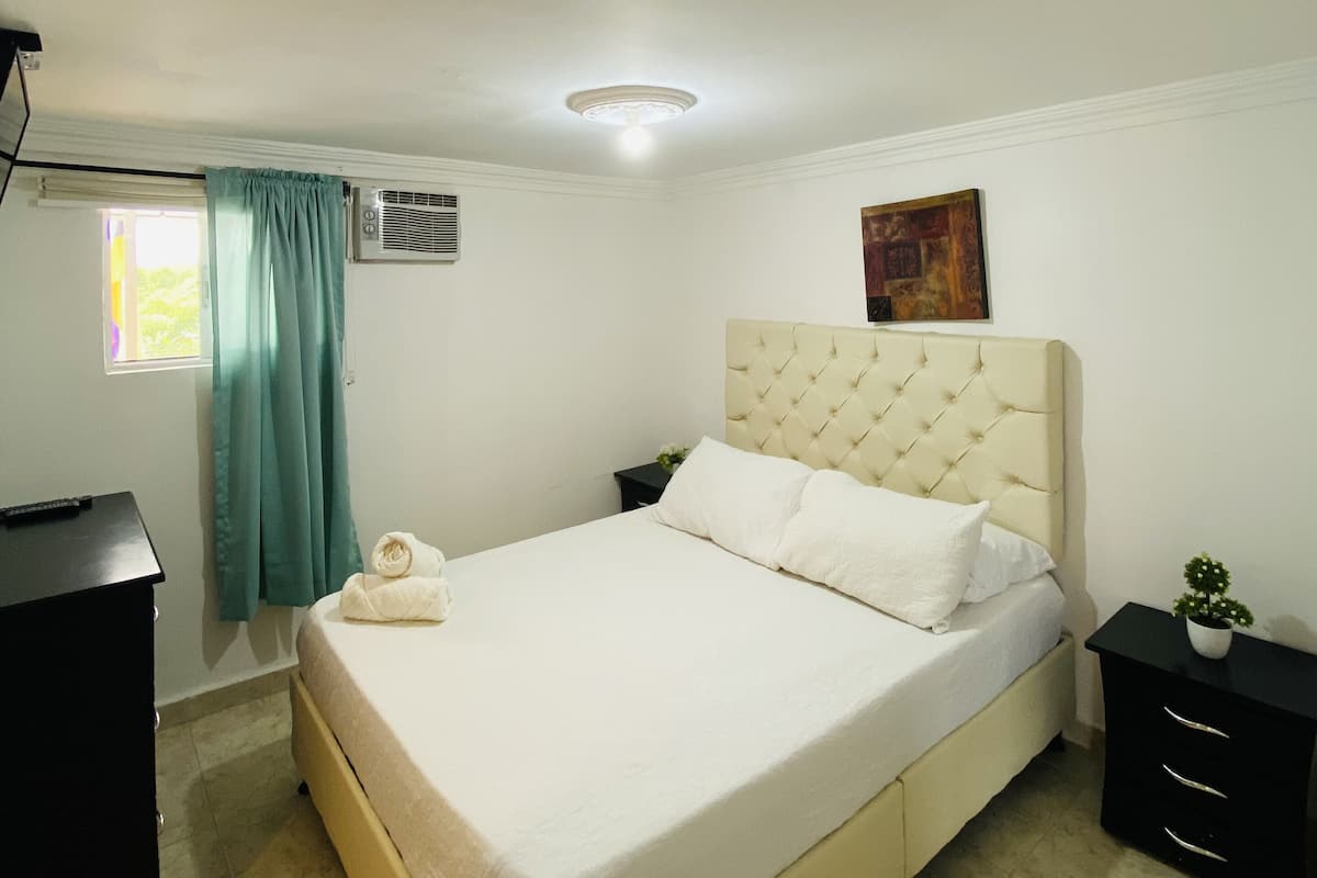 A bedroom with a bed, a nightstand, a small table with a plant, and a window with curtains.