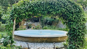 Un chemin de pierres mène à une piscine circulaire surmontée d’une structure en bois, elle-même partiellement recouverte d’une arche de vigne.