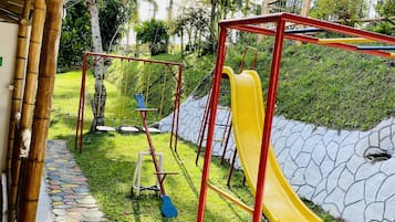 A playground with a yellow slide and red frame, surrounded by greenery and a stone pathway.
