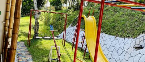 Un parque infantil con un tobogán amarillo y estructura roja, rodeado de follaje y un camino de piedra.