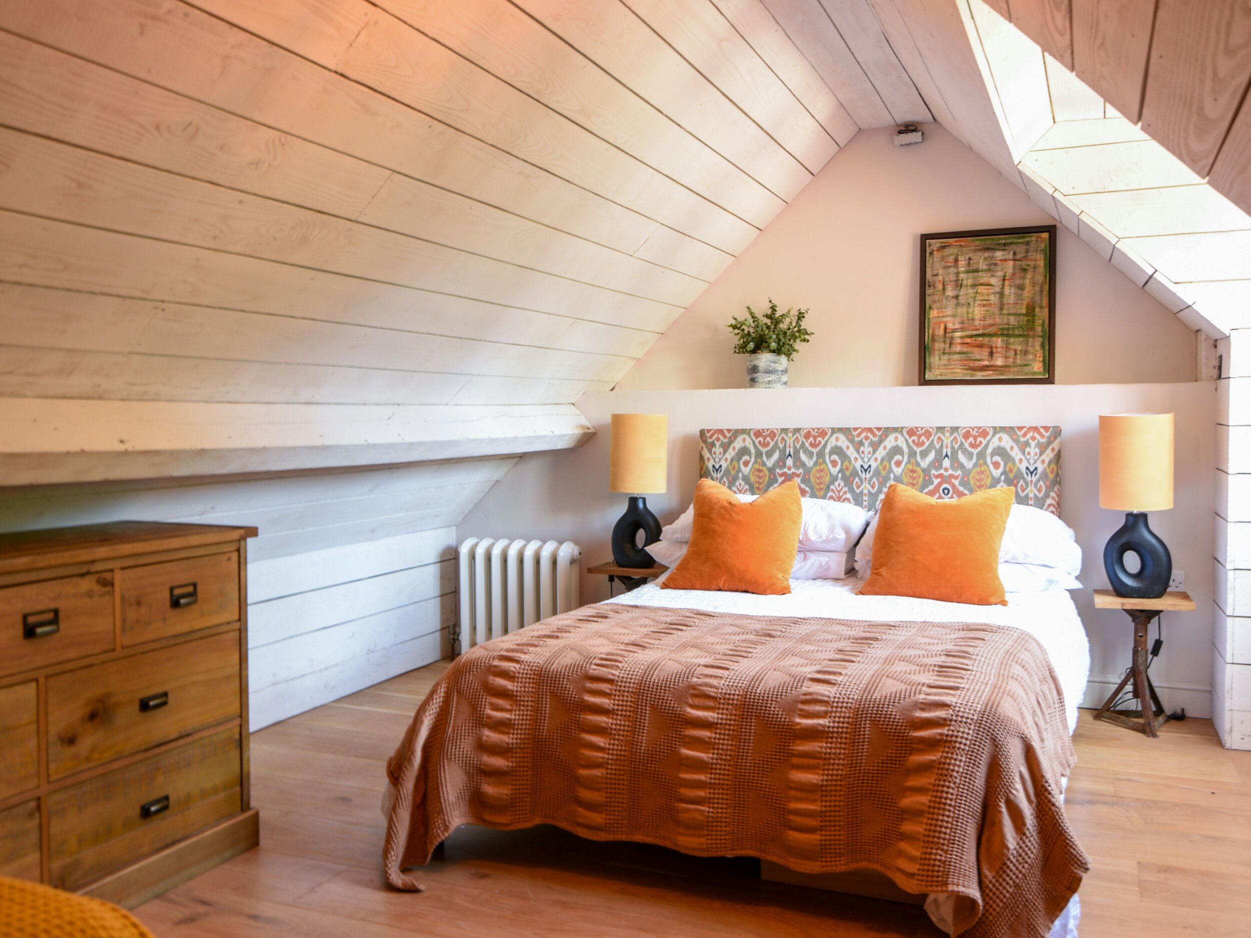 A bedroom with a sloped ceiling, a bed with orange pillows, two bedside lamps, a radiator, a wooden dresser, and a painting on the wall.
