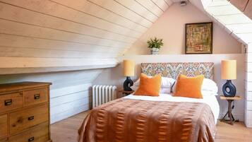 A bedroom with a sloped ceiling, a bed with orange pillows, two bedside lamps, a radiator, a wooden dresser, and a painting on the wall.