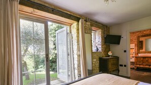 A bedroom with a large bed, a stone wall, a TV mounted on the wall, and a view of the outdoors. - The Carriage House (Lyme Regis)