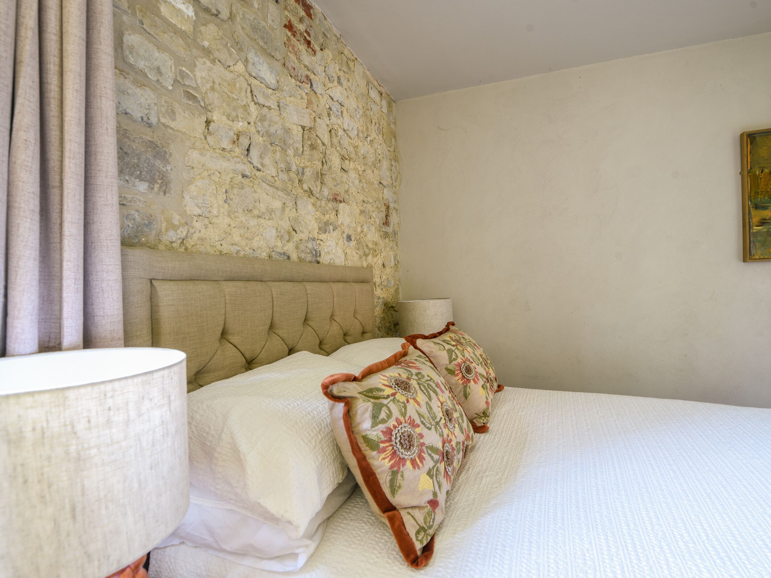 A bedroom with a stone wall, a bed with white bedding and floral pillows, a bedside lamp, and a painting on the wall.