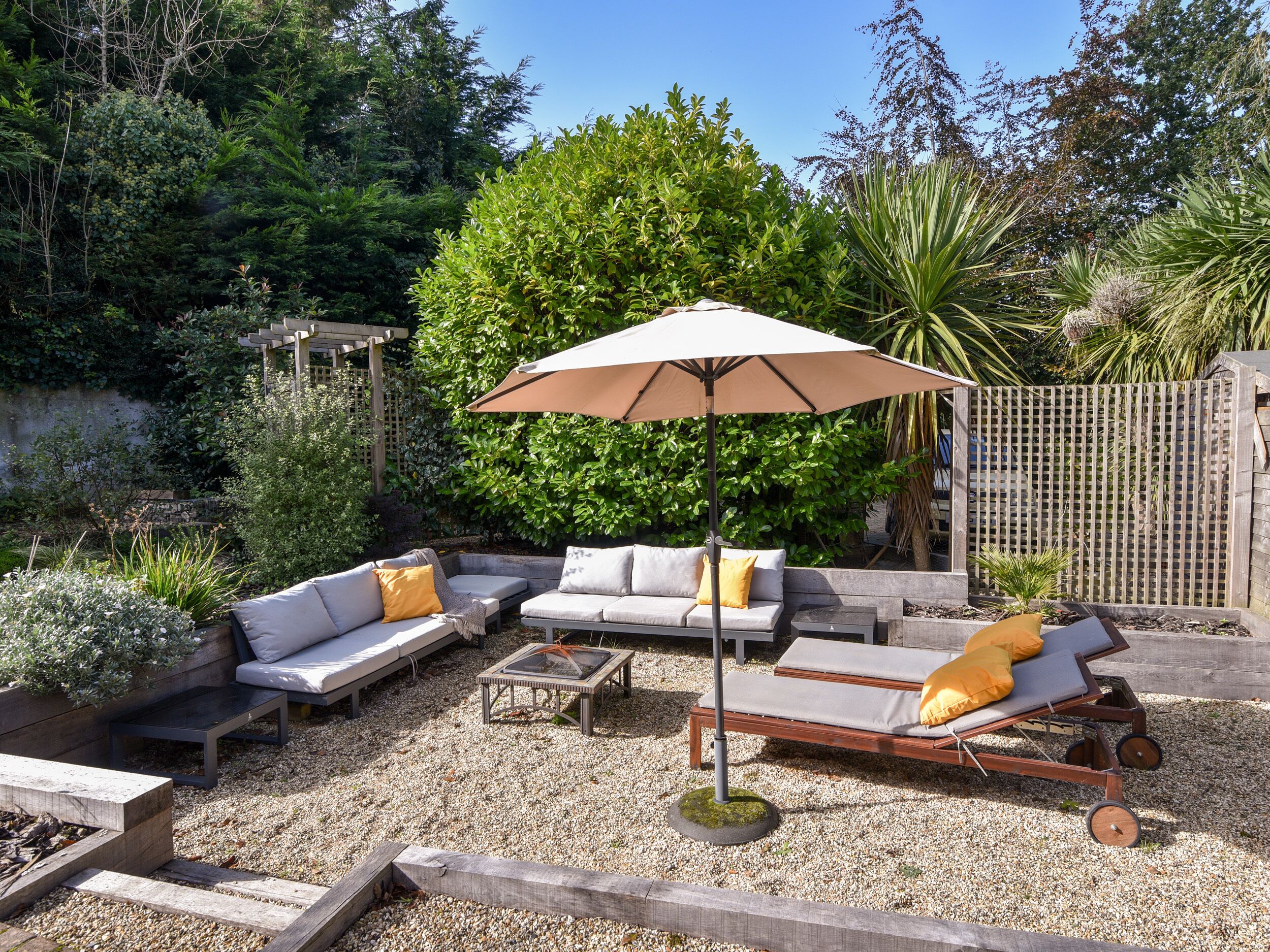 A patio area with a seating arrangement, including a sofa, a lounge chair, and a table. There is a pergola structure and a wooden fence in the background.