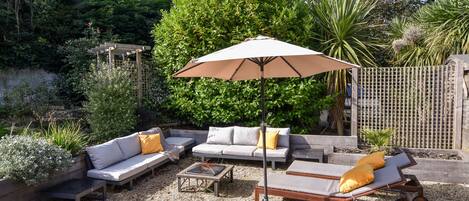 A patio area with a seating arrangement, including a sofa, a lounge chair, and a table. There is a pergola structure and a wooden fence in the background.