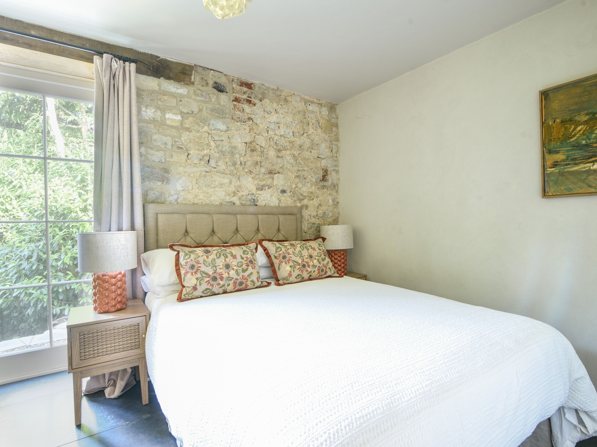A bedroom with a stone wall, a bed with floral pillows, a nightstand with a lamp, and a window with curtains.