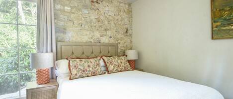 A bedroom with a stone wall, a bed with floral pillows, a nightstand with a lamp, and a window with curtains.