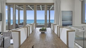 A spacious dining area with a wooden table, white cushioned chairs, a central plant, and a view of the ocean through large glass doors.