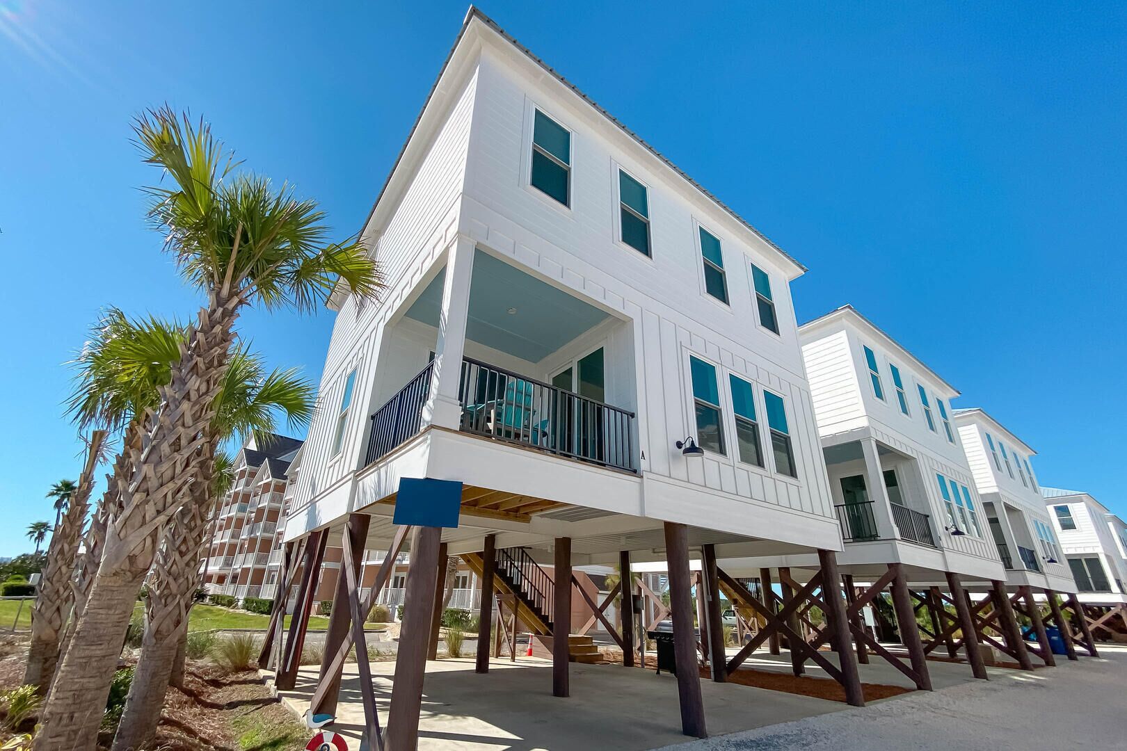 A multi-story beachfront house with a balcony, wooden deck supports, and palm trees.