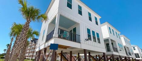 A multi-story beachfront house with a balcony, wooden deck supports, and palm trees.