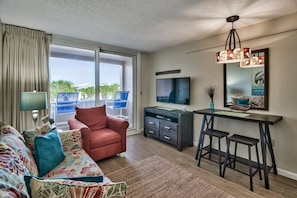 A living room with a sofa, a red armchair, a TV, a desk with stools, and a large window with a view of palm trees. - Jetty View at Jetty East! One FREE Beach Chair Setup Each Day of Your Stay! (Destin)