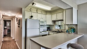 A kitchen with white cabinets, a refrigerator, a microwave, a stove, and a sink. There are two stools at the island. - Jetty View at Jetty East! One FREE Beach Chair Setup Each Day of Your Stay! (Destin)