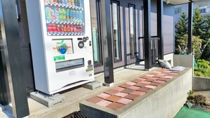 A vending machine with various drink options, a concrete step leading to a building entrance, and a small garden area with a green mat and some plants. - Sho inn MINIMAL HOTEL (Otaru)