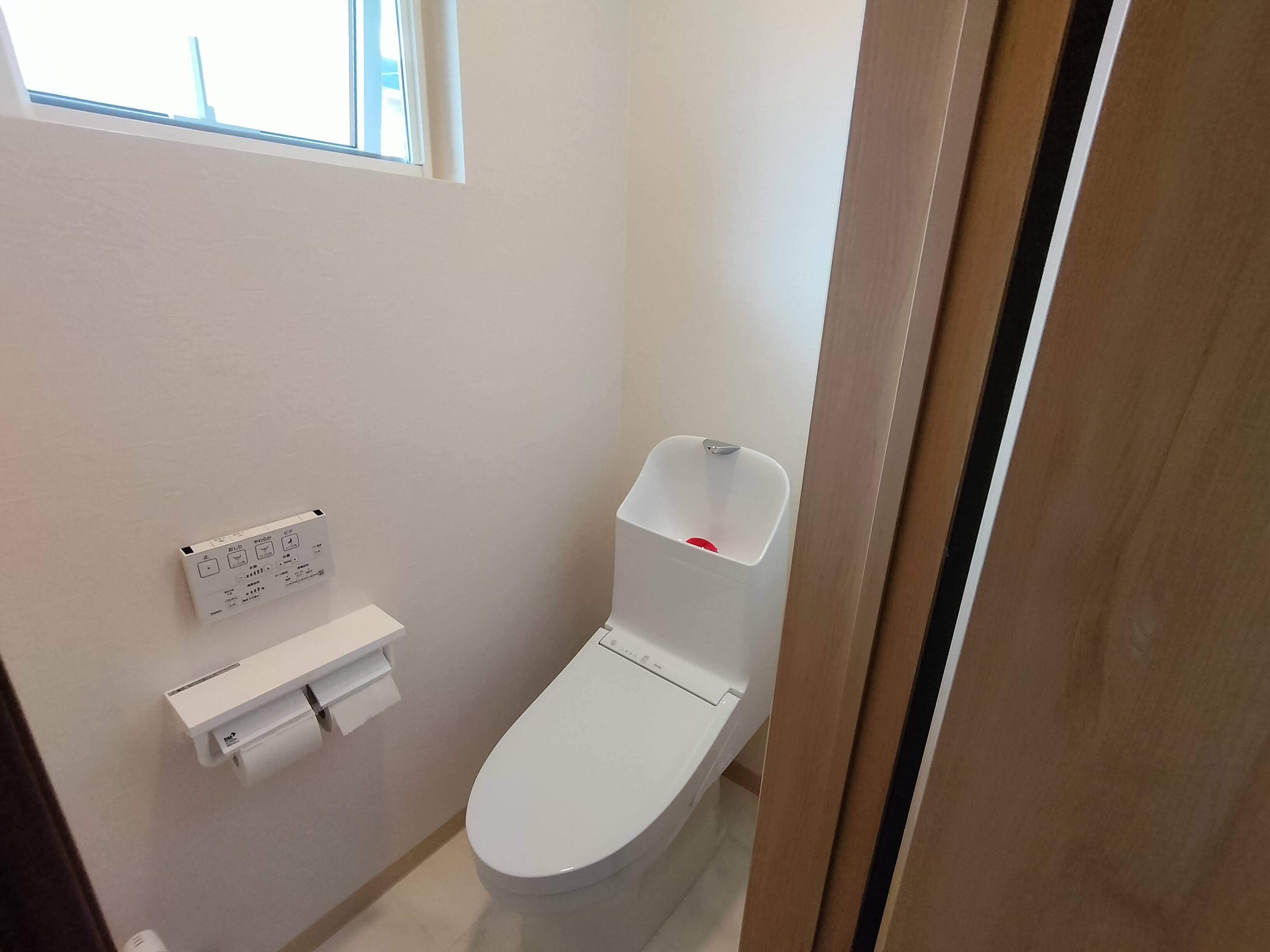 A small, clean bathroom with a toilet, a white toilet paper holder, and a window above the toilet.