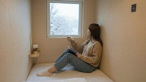 A person sitting on a bed in a small room with a window, a shelf with a coffee cup, and a thermostat on the wall. - Sho inn MINIMAL HOTEL (Otaru)