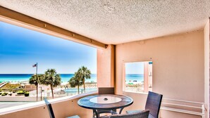 Een balkon met een ronde tafel en vier stoelen, met uitzicht op een strand met palmbomen en een helderblauwe lucht.
