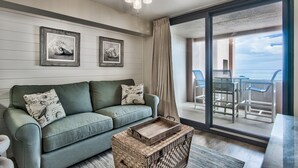 Ein Wohnzimmer mit einem grünen Sofa, zwei gerahmten Bildern, einem Deckenventilator und Blick auf einen Balkon mit Gartenmöbeln.