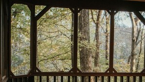 Um deck de madeira com um banco e uma mesa, com vista para uma área arborizada através de grandes janelas.