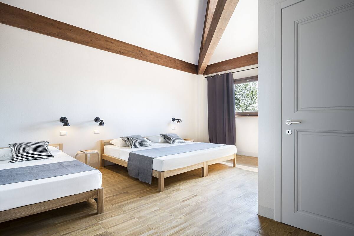 A bedroom with two beds, wooden beams, a white door, and a window with curtains.
