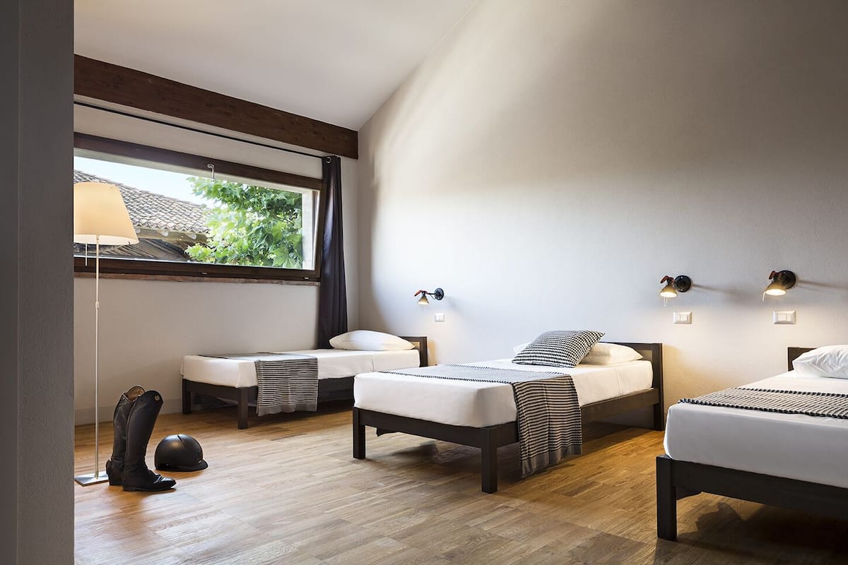 A modern bedroom with two single beds, a standing lamp, and a large window with a view of greenery.