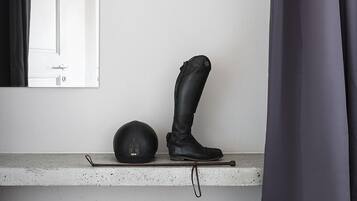 Una habitación con piso de madera, pared blanca, ventana con cortina negra, un cuadro colgado, un casco negro y un par de botas negras sobre una repisa.