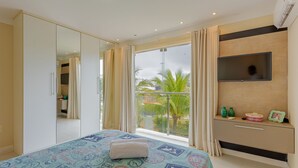 Habitación de hotel con una cama grande, un armario, televisor montado en la pared y un balcón con vistas a palmeras.