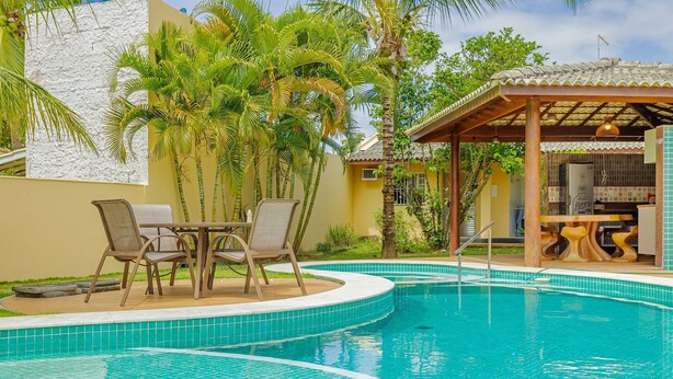 Una piscina con borde de azulejos, rodeada de palmeras y una zona de estar cubierta con sillas.