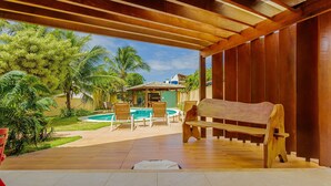 Un patio al aire libre con un banco de madera, una piscina y un área cubierta con vistas a una casa y palmeras.