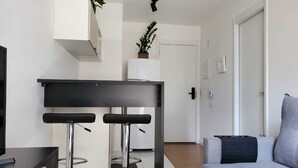 A modern living room with a grey sofa, a black dining table with two bar stools, a TV, and a white wall-mounted shelf with a plant. - Sunflower Stays (São Paulo)