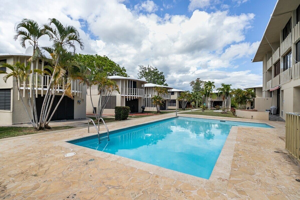 Una piscina rodeada de edificios con balcones, palmeras y un cielo despejado.