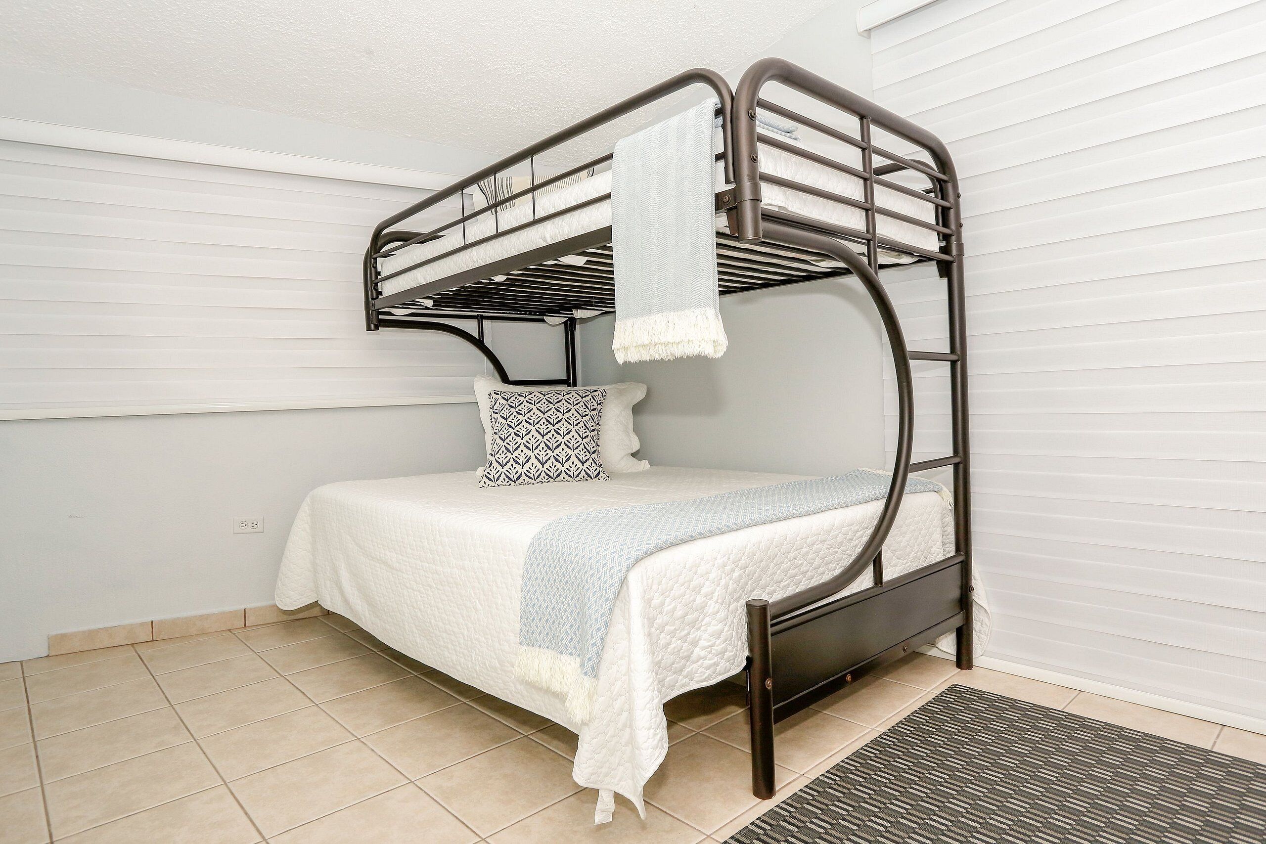A bunk bed with a white mattress and a patterned pillow, a grey blanket, and a dark metal frame.