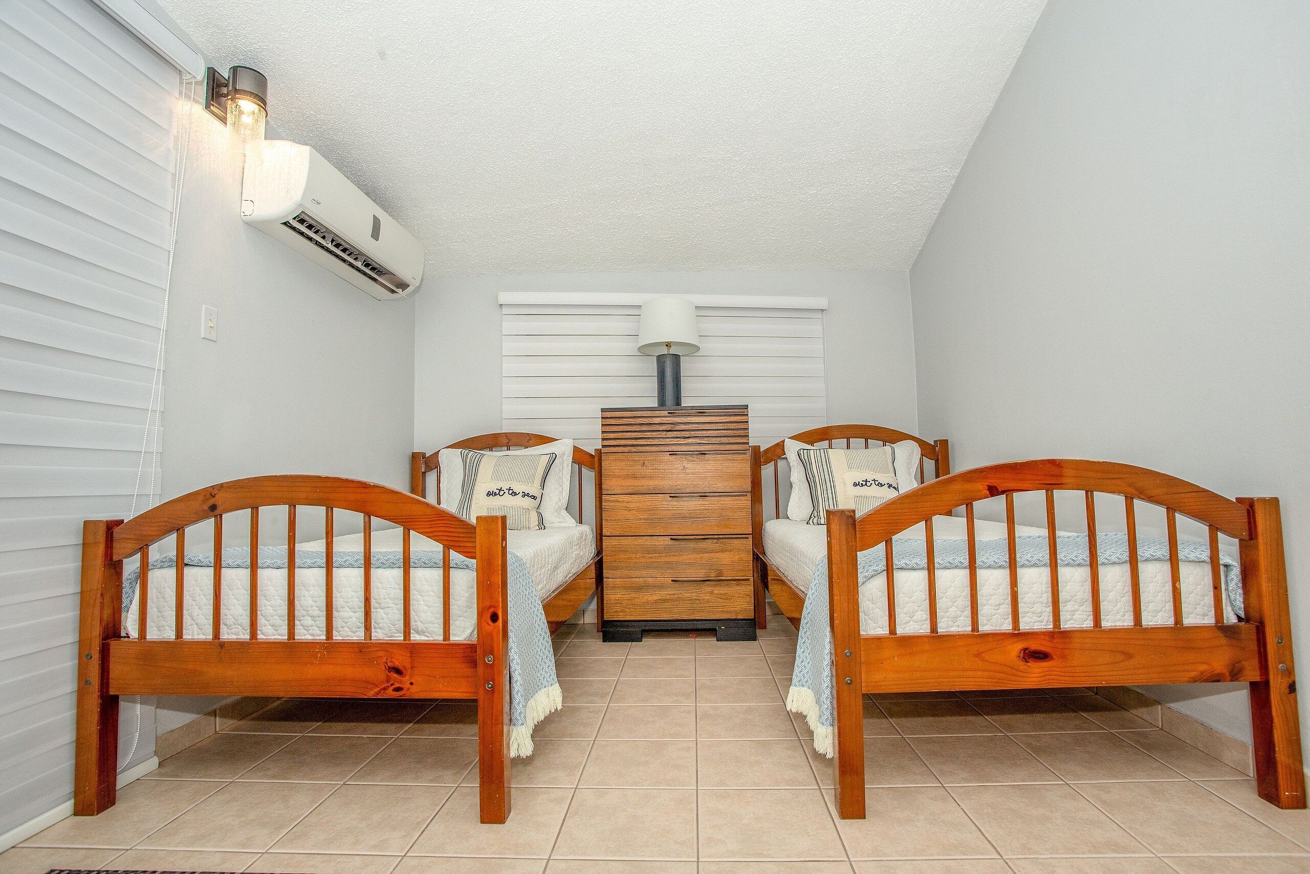 A bedroom with two wooden beds, a central dresser, and an air conditioning unit.