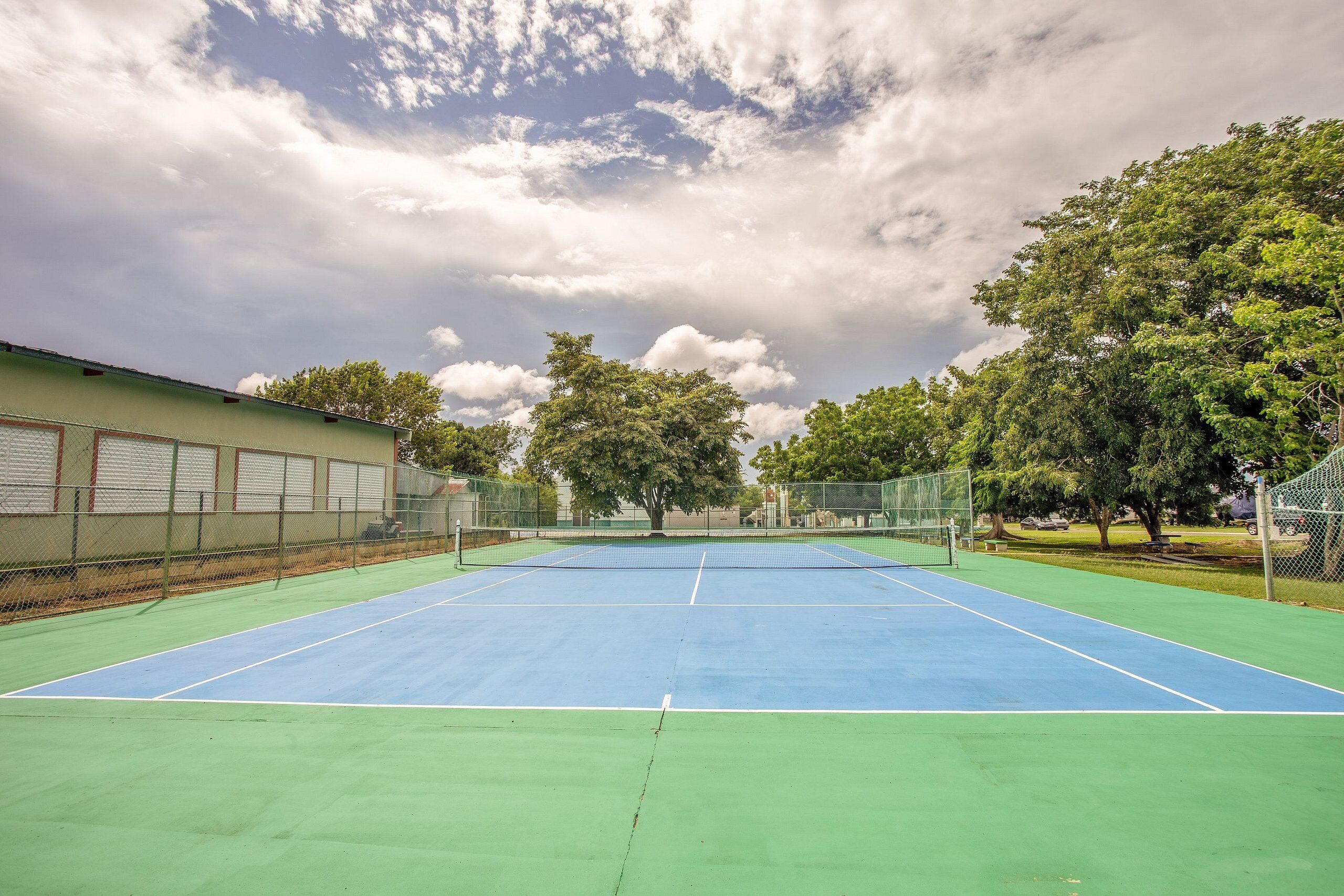 Una cancha de tenis al aire libre con superficie verde y azul, rodeada de árboles y una cerca.