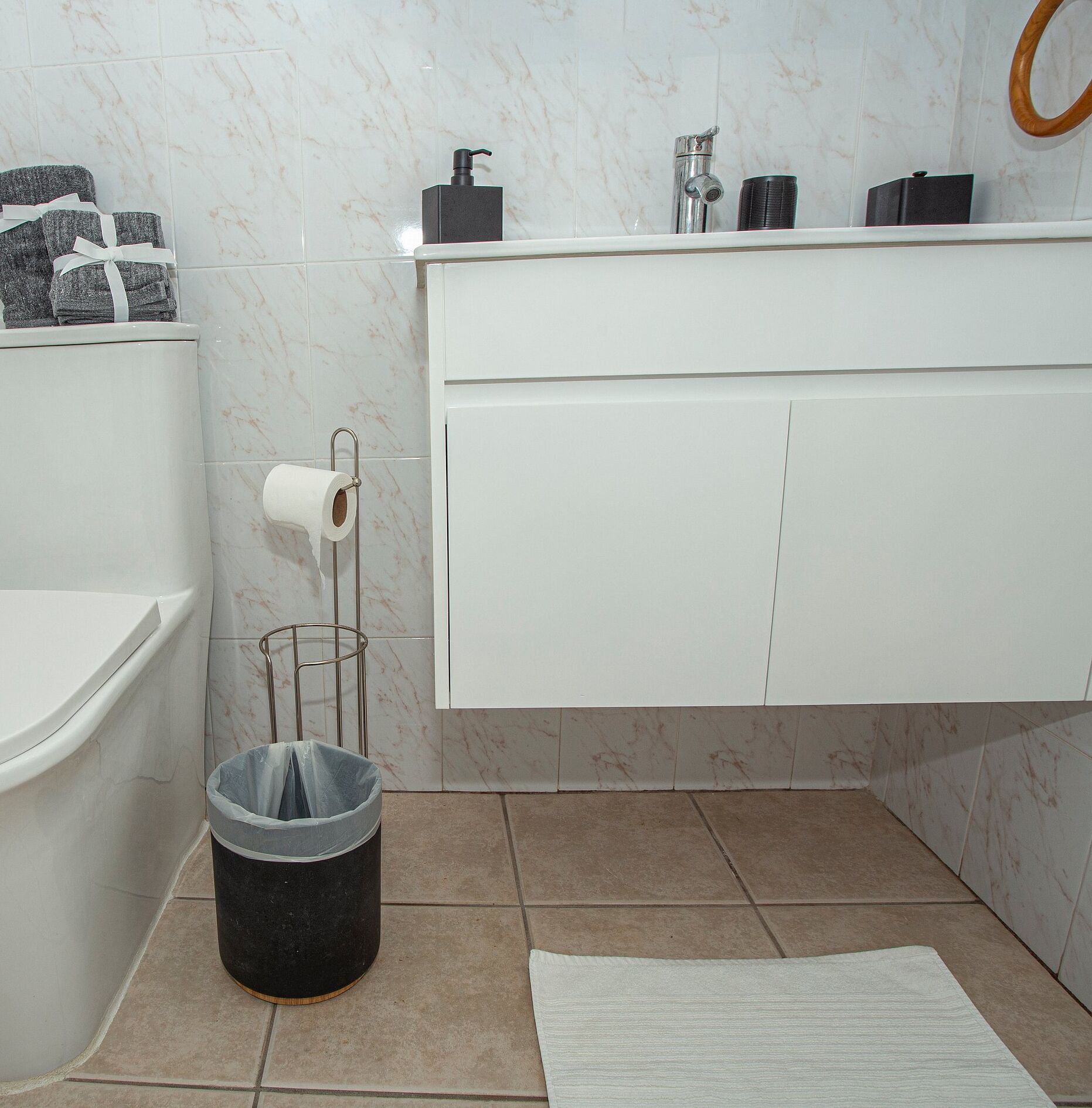 A bathroom with a toilet, a sink, a mirror, a trash bin, and a roll of toilet paper.