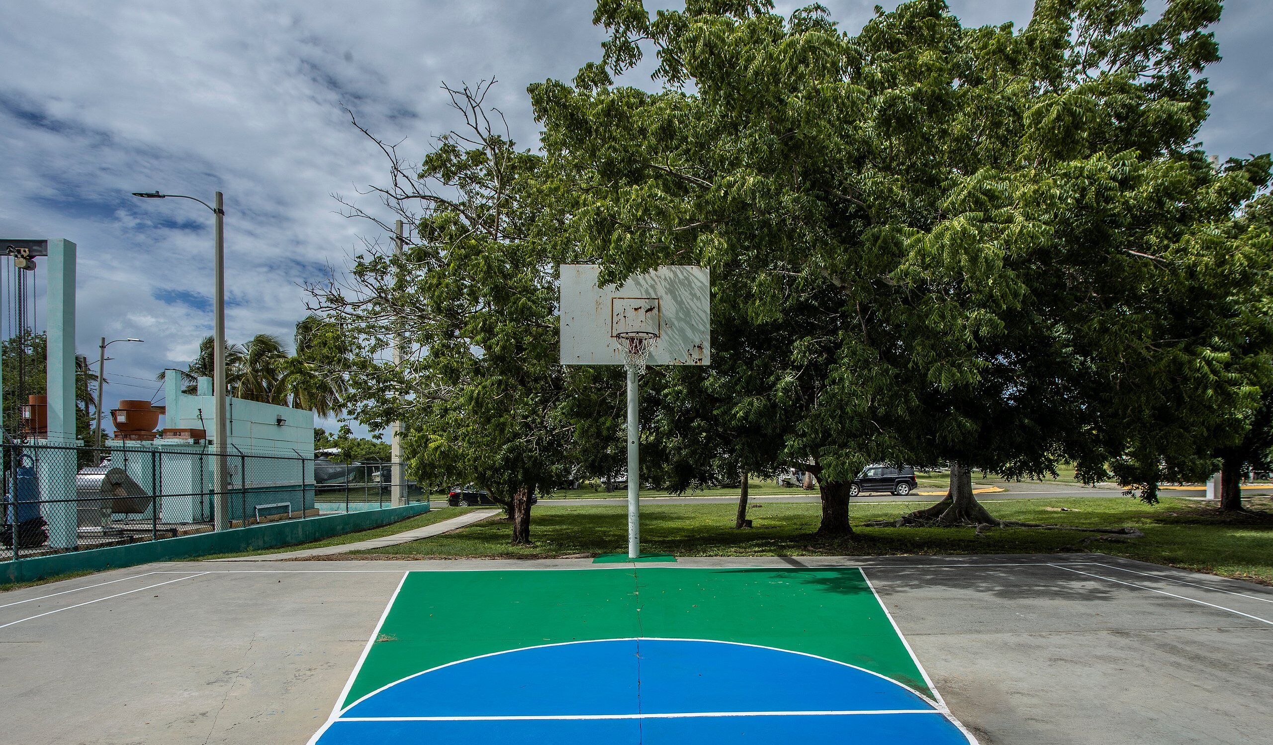 An outdoor basketball court with a green and blue playing surface, surrounded by trees and a fence.