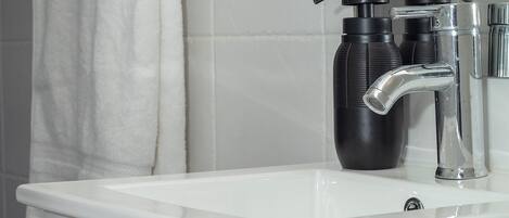 Casa de banho com lavatório branco, toalha pendurada num porta-toalhas e dois dispensadores de sabonete pretos.