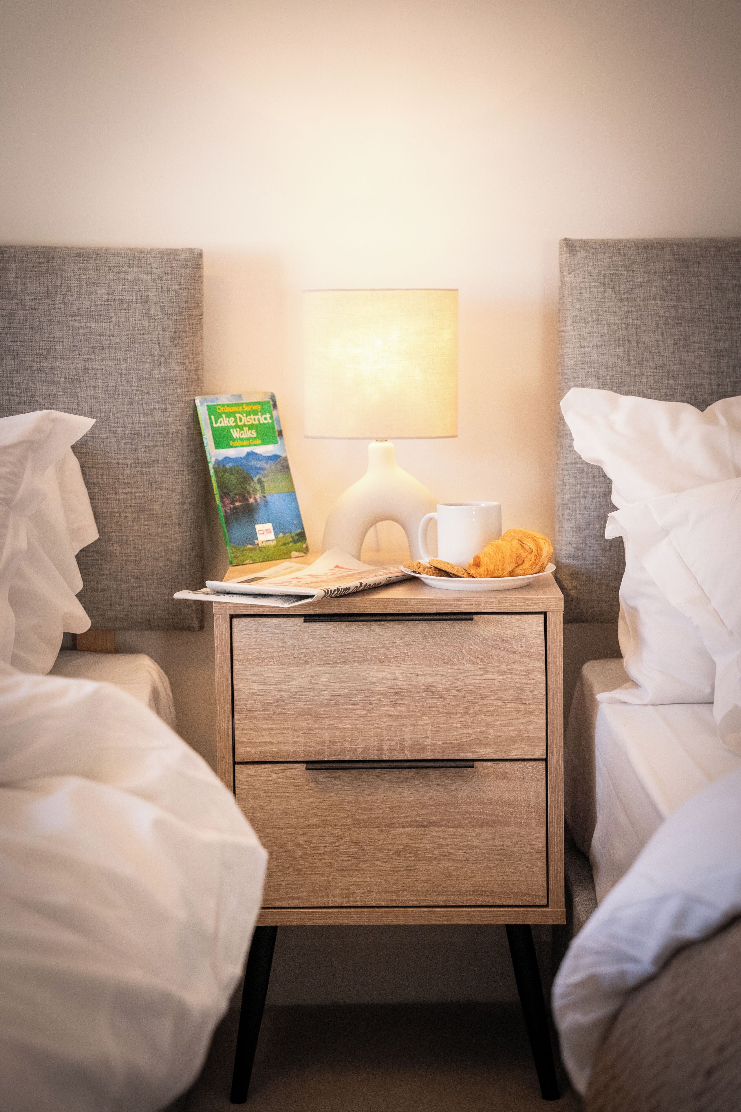 Ein ordentlich gemachtes Bett mit weißen Leinen, ein Nachttisch mit Lampe, einer Broschüre, einer Tasse und einem Croissant.