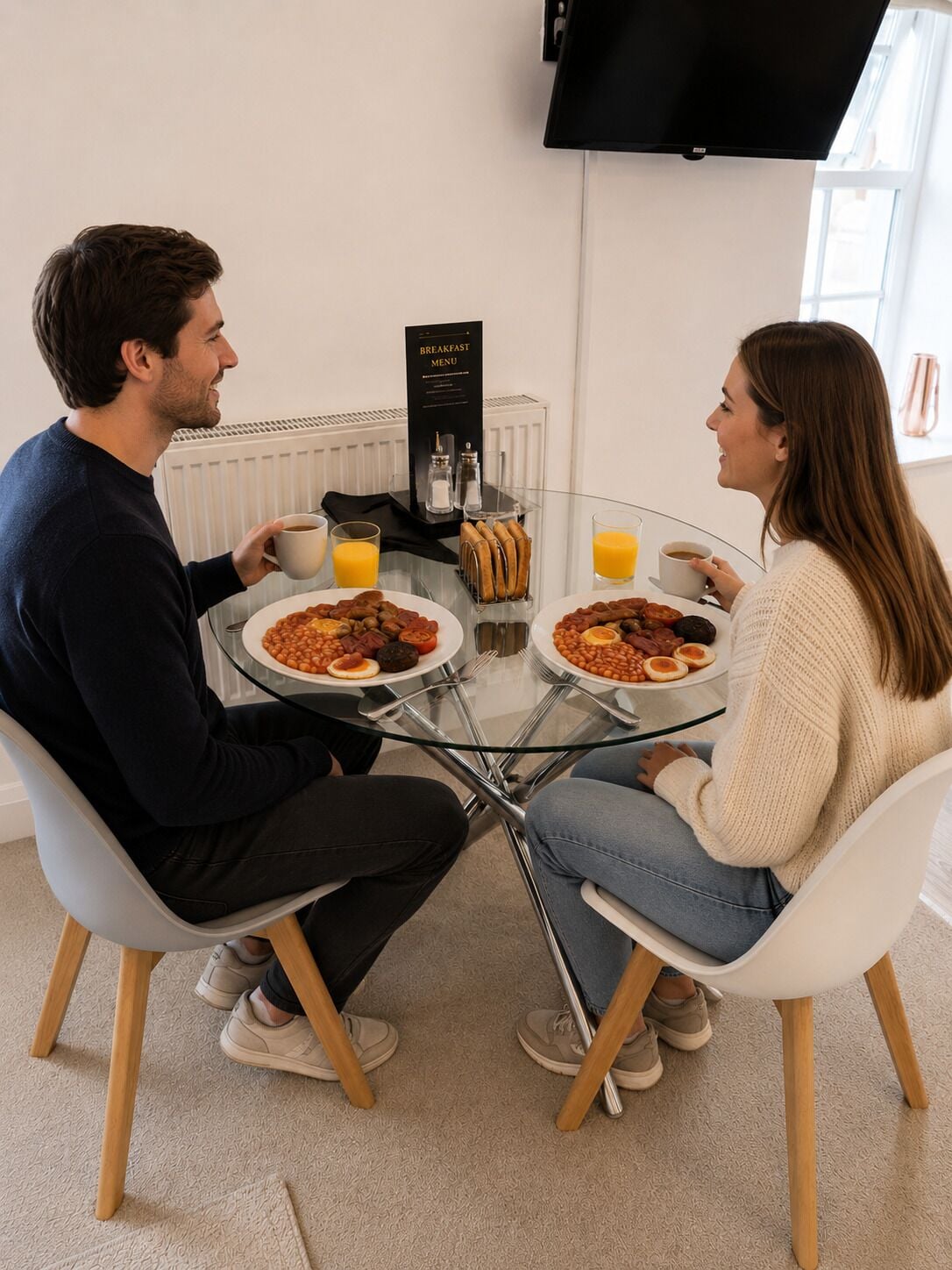 Two people seated at a glass table with plates of food, a breakfast menu stand, and a TV mounted on the wall.
