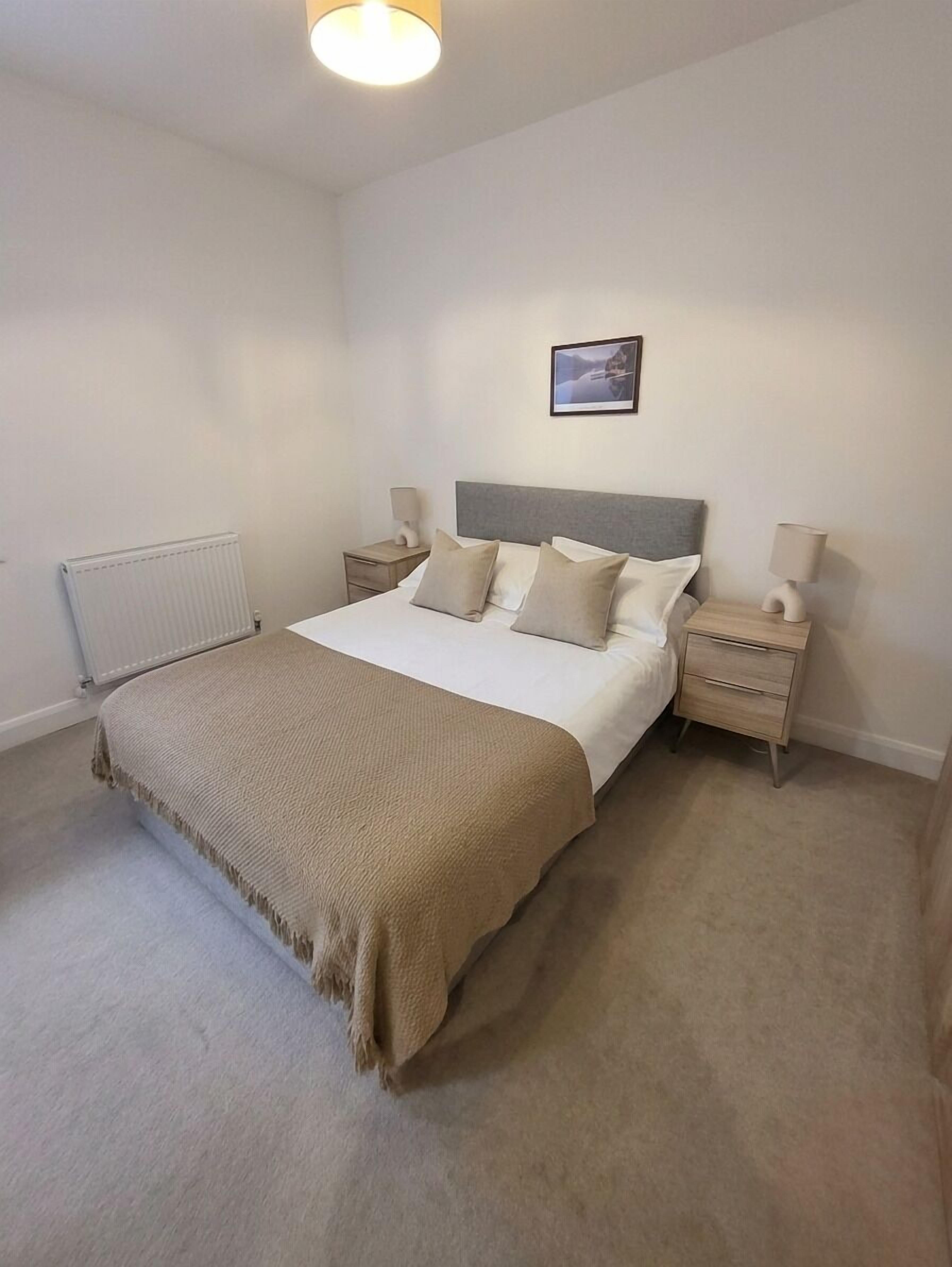 A bedroom with a bed, two bedside tables, a radiator, a picture on the wall, and a carpeted floor.