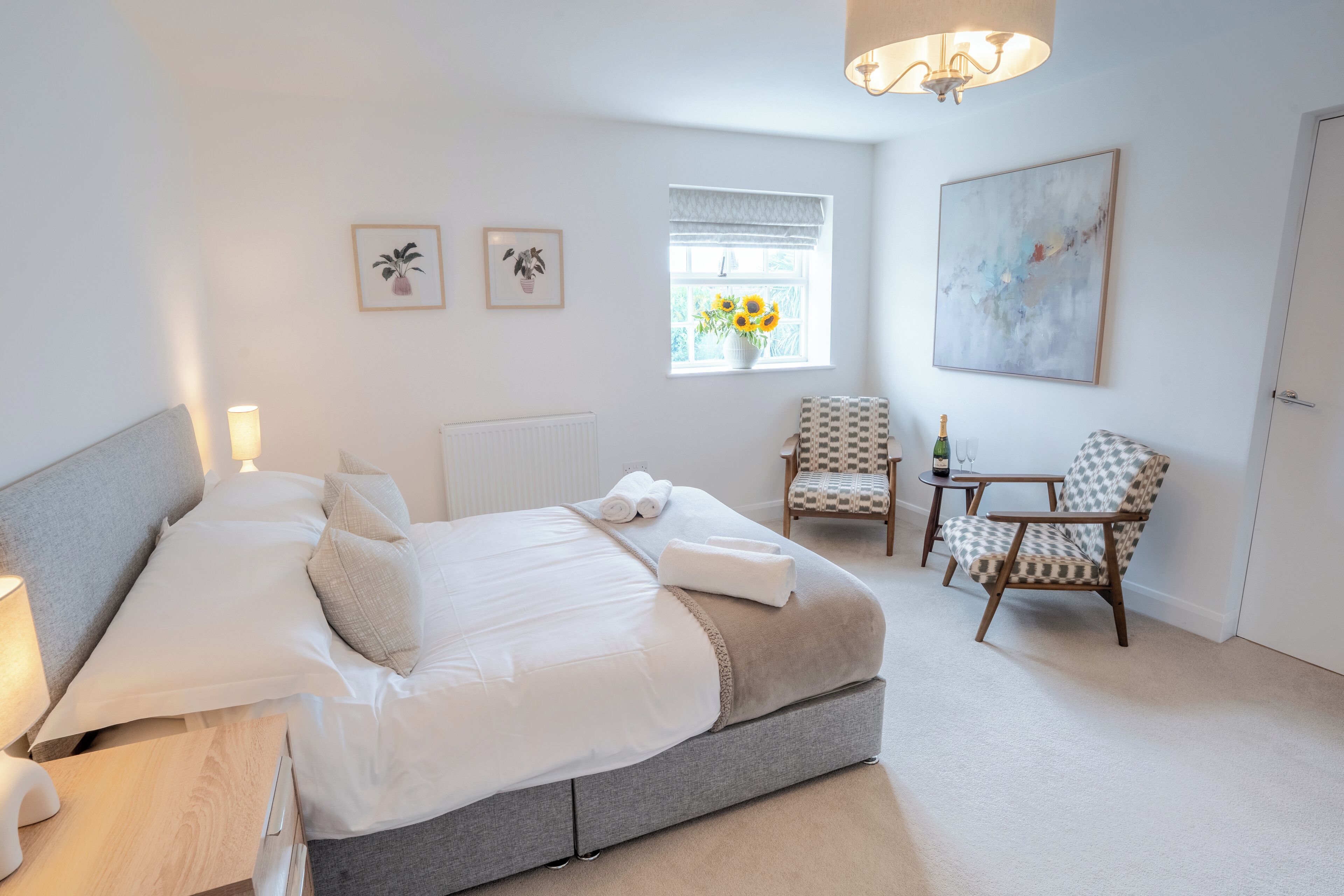A bedroom with a large bed, bedside tables, a chair, a small table, a window with sunflowers, and a painting on the wall.