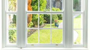 Ein großes, rundes Fenster mit vielen Glasscheiben, das einen Blick auf einen üppigen Garten und ein Steinhaus freigibt.