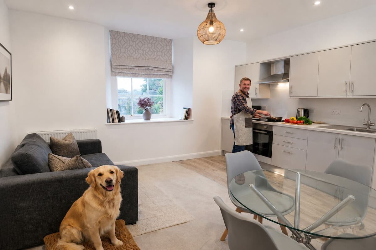 Une cuisine moderne avec des meubles blancs, un coin repas doté d’une table en verre et un salon avec un canapé gris et un chien.