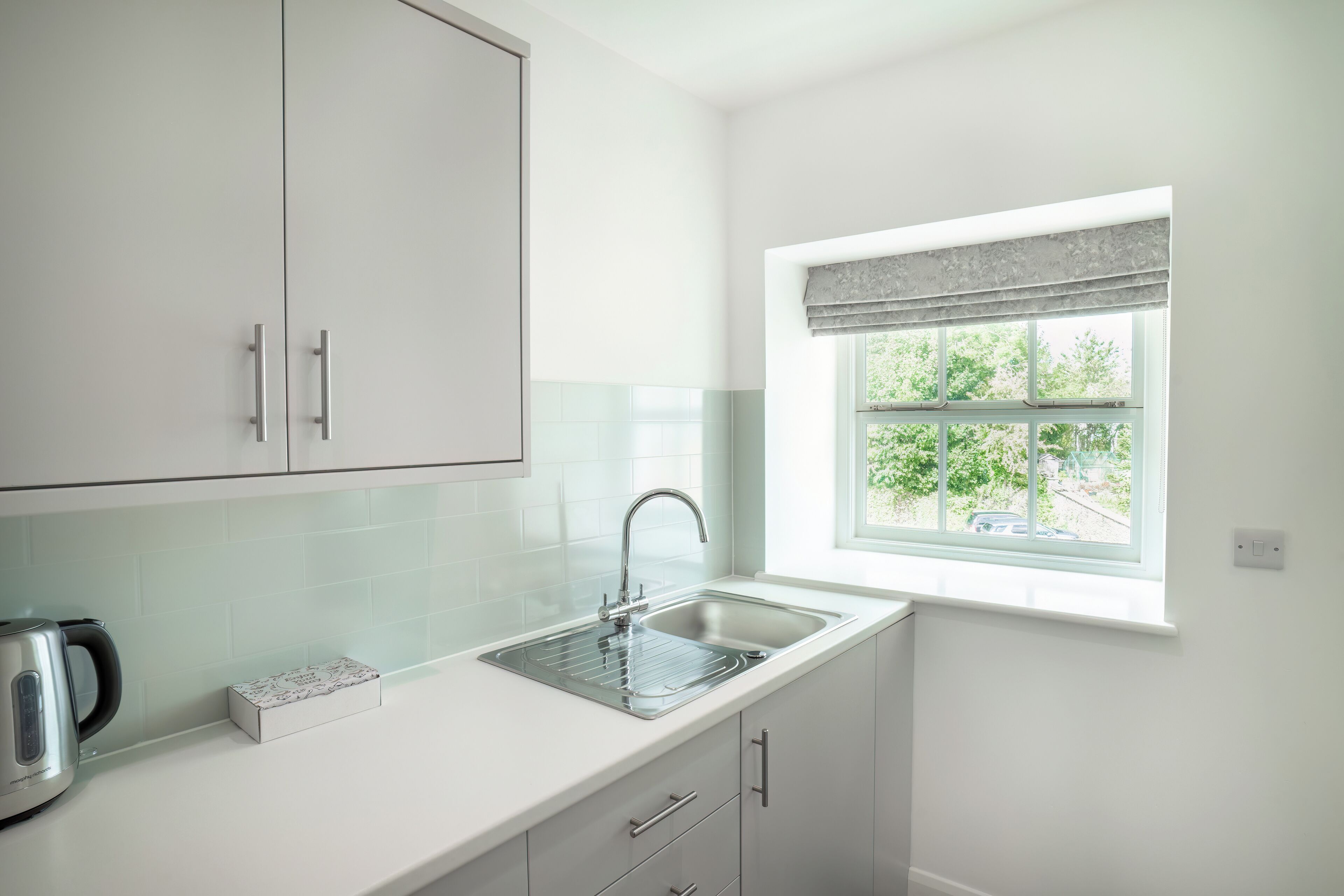 Une cuisine moderne avec des meubles blancs, un évier en acier inoxydable et une fenêtre donnant sur un paysage verdoyant.